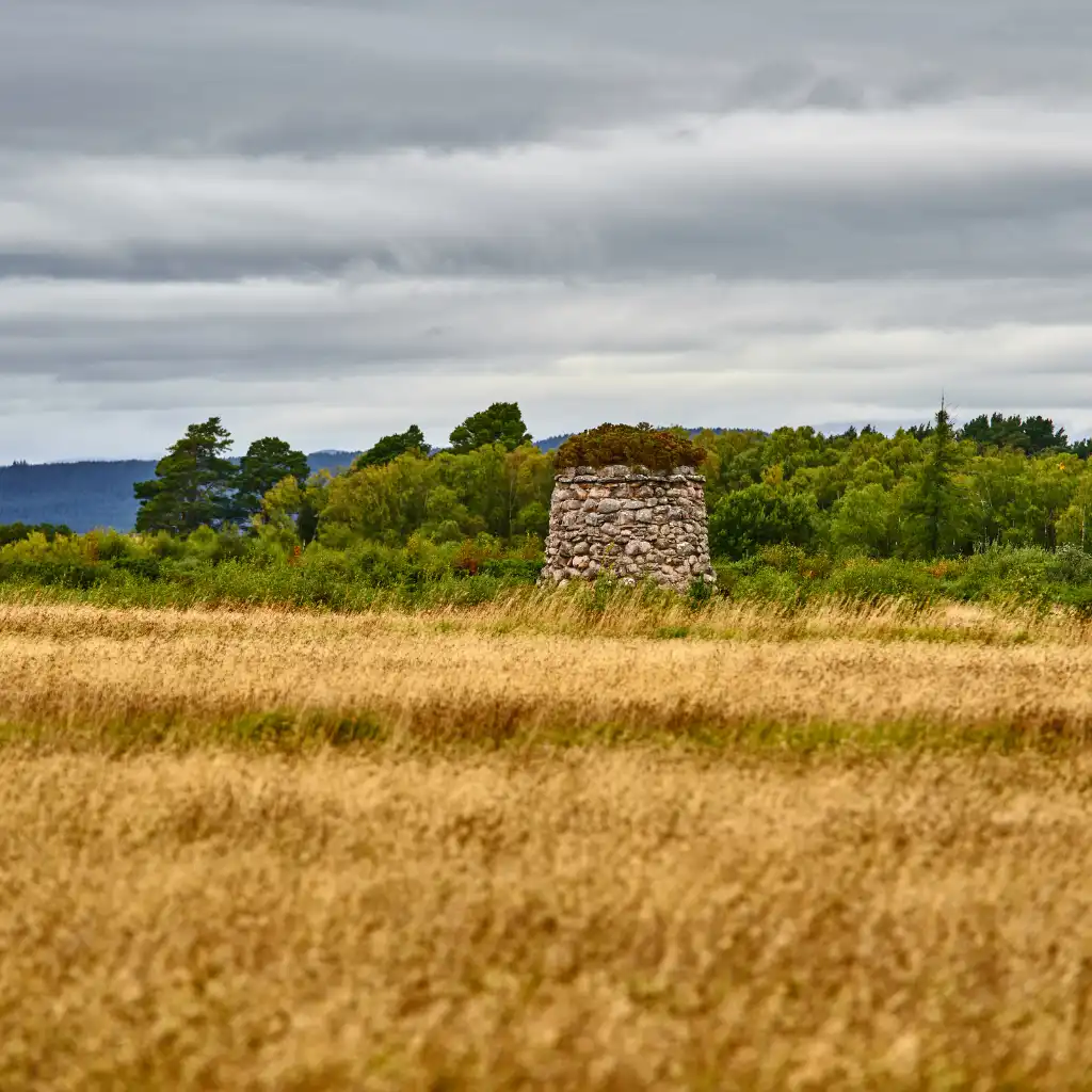 Culloden, Scotland battlefield stone cairn amid golden fields with green forest and hills under cloudy sky