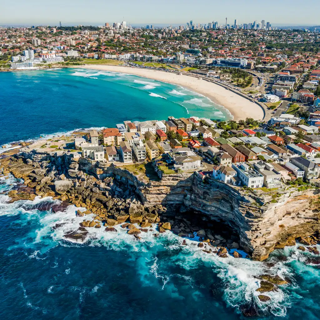 Bondi Beach Sydney aerial view of turquoise waves, rocky cliffs and curving sandy shoreline with city skyline