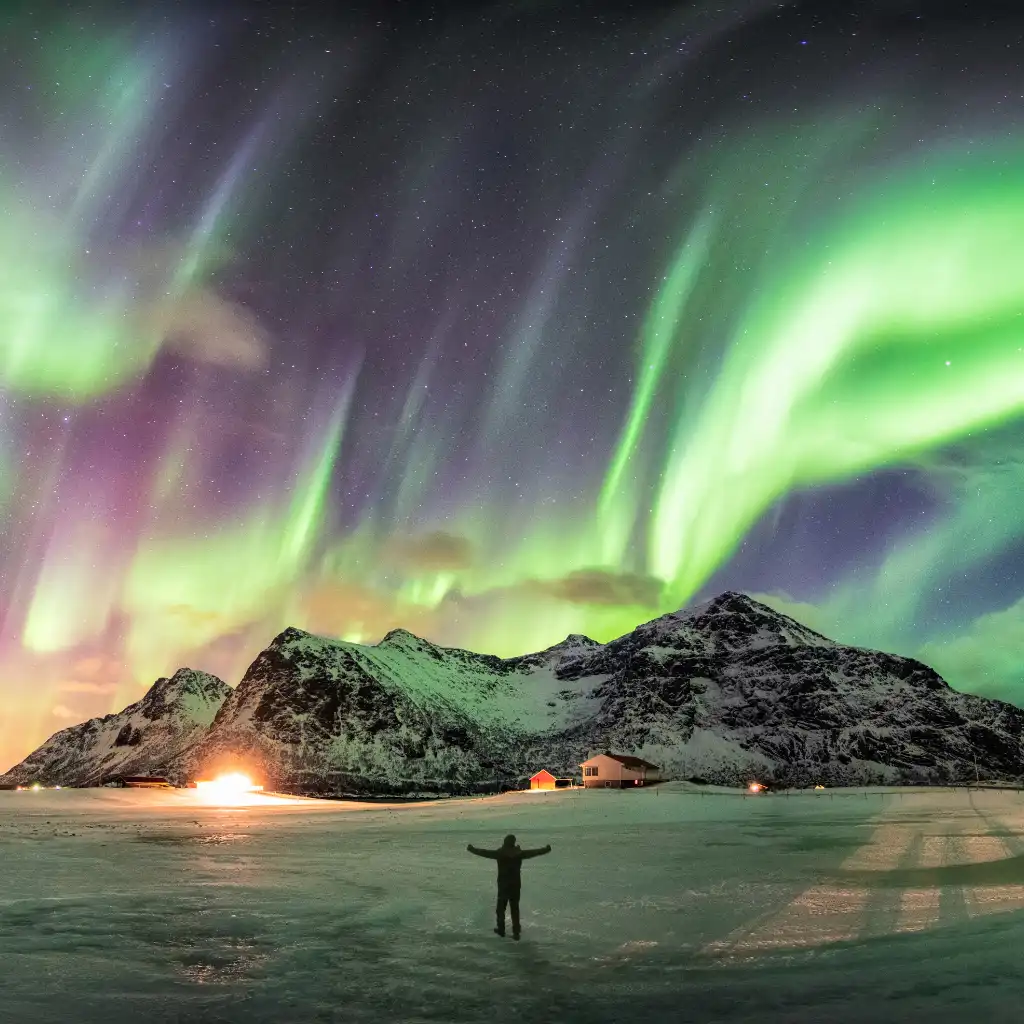 Northern Lights Norway over snowy mountains with person under green aurora on frozen lake at night