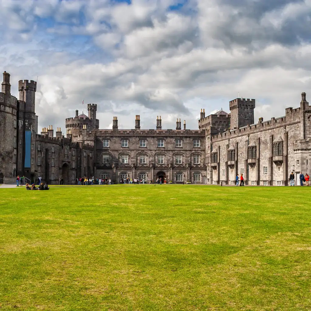Kilkenny Castle stone towers and facade above green lawn with visitors on a sunny day in Ireland