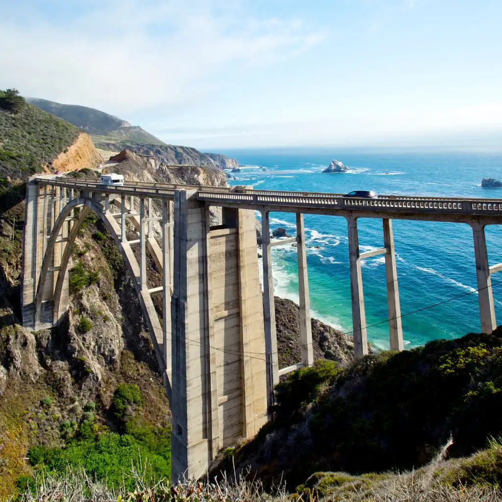 Highway 1 bridge in Big Sur California with ocean cliffs turquoise water and passing vehicles