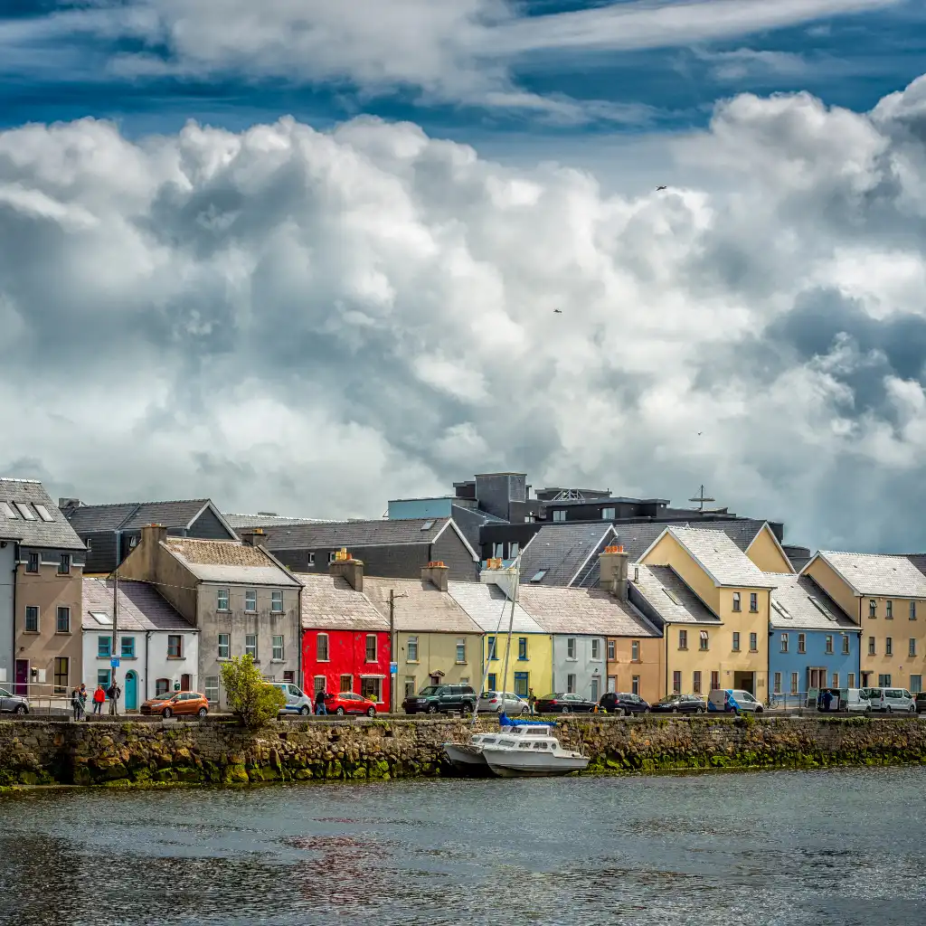 Colorful houses along Galway waterfront with boats and cars under dramatic clouds on a cloudy morning