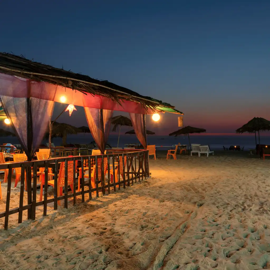 Goa, India beach shack with thatched roof, lights and seating on sandy shore at dusk